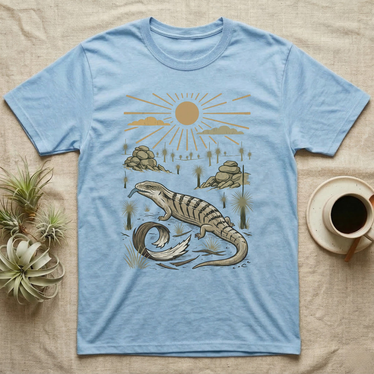 Blue-Tongued Skink Home Range T-Shirt in Light Blue — Woodcut-style illustration of a blue-tongued skink walking across bare soil with its tongue out, shed skin and grass trees behind, no on-shirt text.