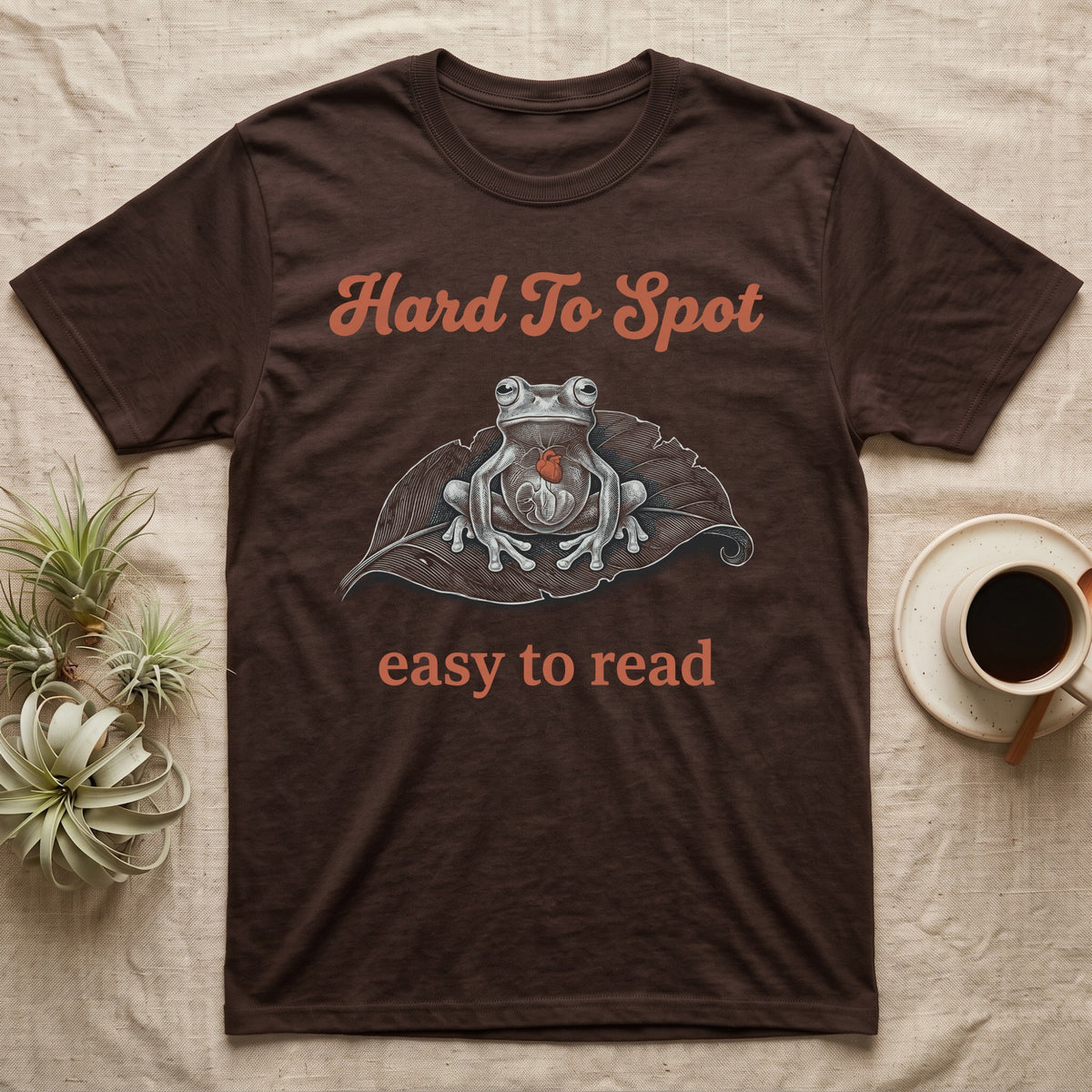 Glass Frog Engraving T-Shirt in Dark Chocolate — White-line engraving of a glass frog sitting on a leaf with a coral heart showing through its belly, between Hard To Spot and easy to read