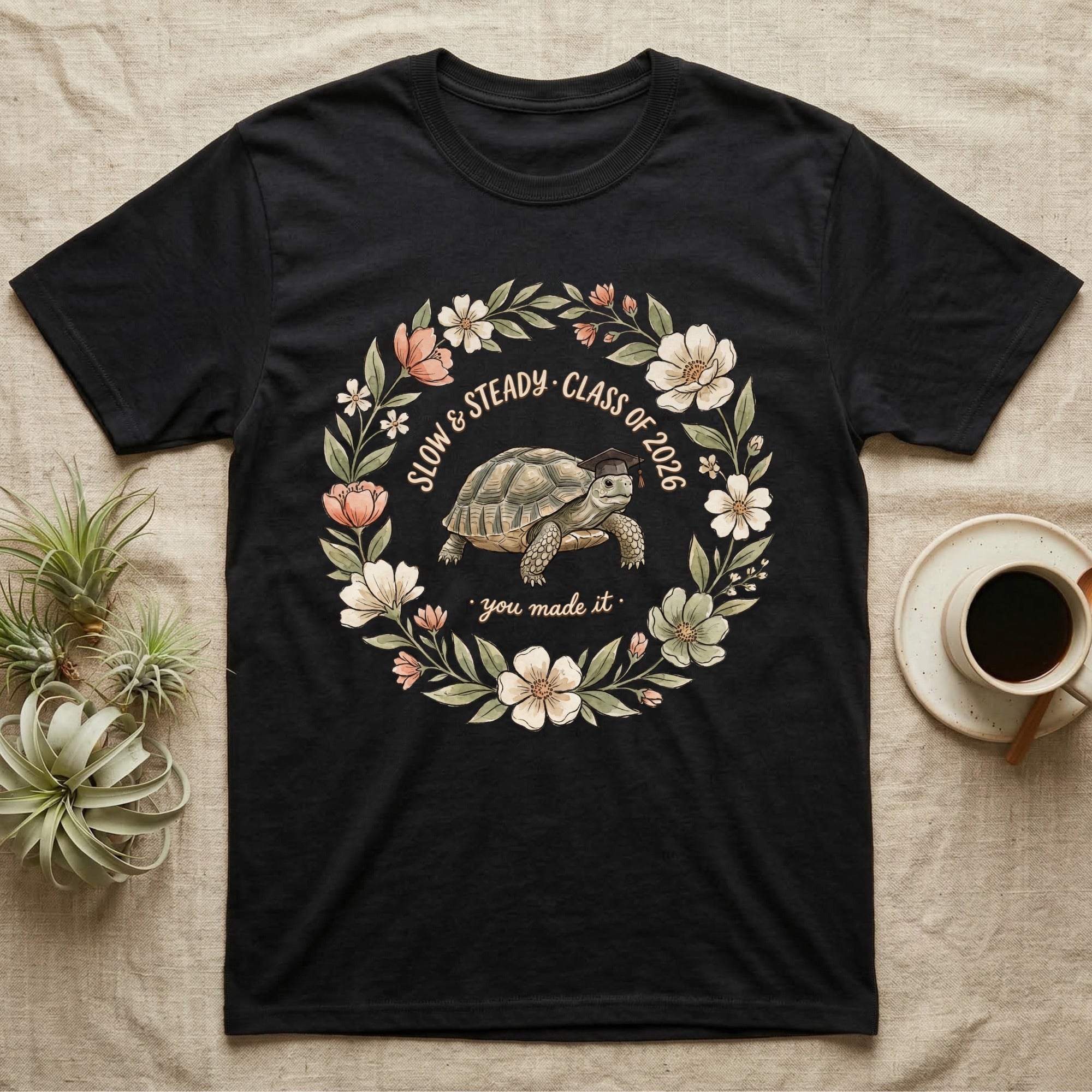 Slow And Steady Graduation T-Shirt in Black — Watercolour illustration of a tortoise in a graduation cap inside a floral wreath with "Slow & Steady, Class of 2026."