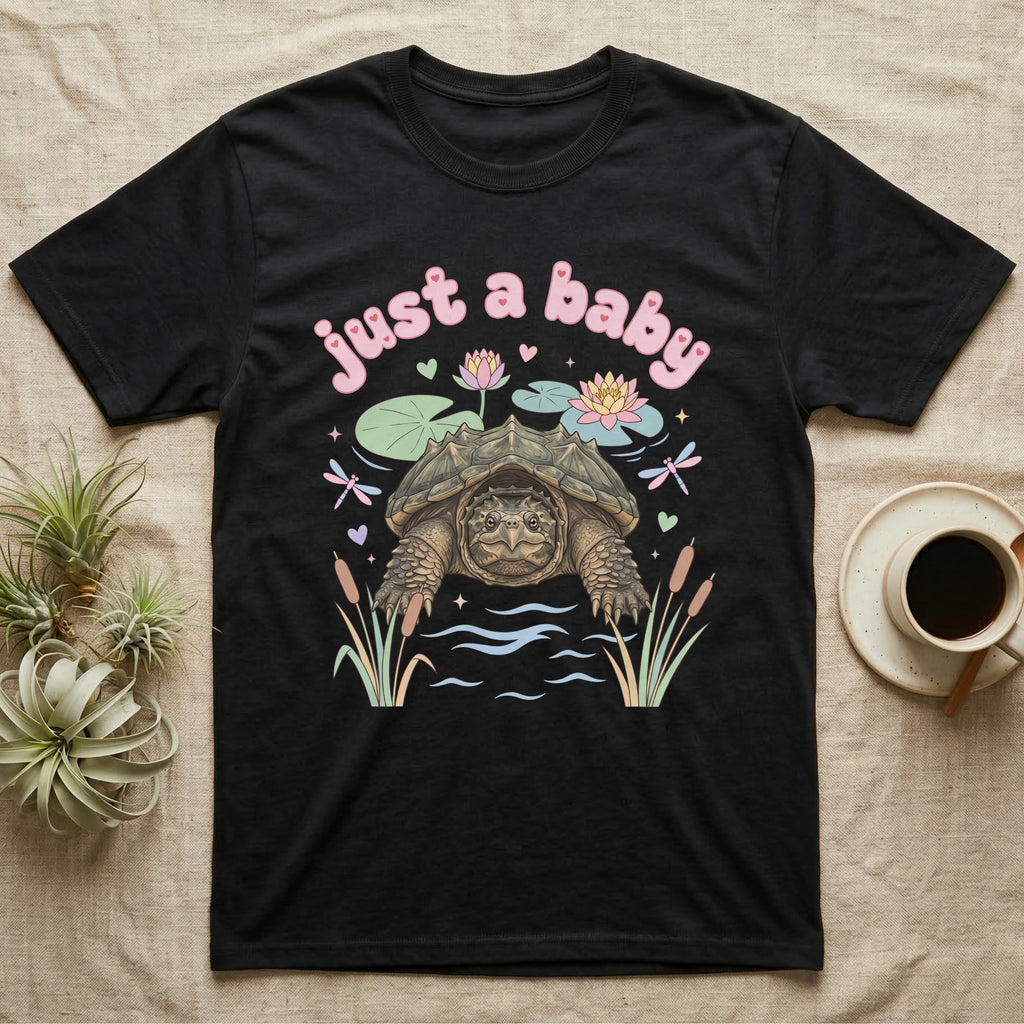 Common Snapping Turtle Baby T-Shirt in Black — Snapping turtle with hooked beak and spiky shell among pastel lily pads under puffy pink bubble text "just a baby".