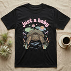 Common Snapping Turtle Baby T-Shirt in Black — Snapping turtle with hooked beak and spiky shell among pastel lily pads under puffy pink bubble text "just a baby".