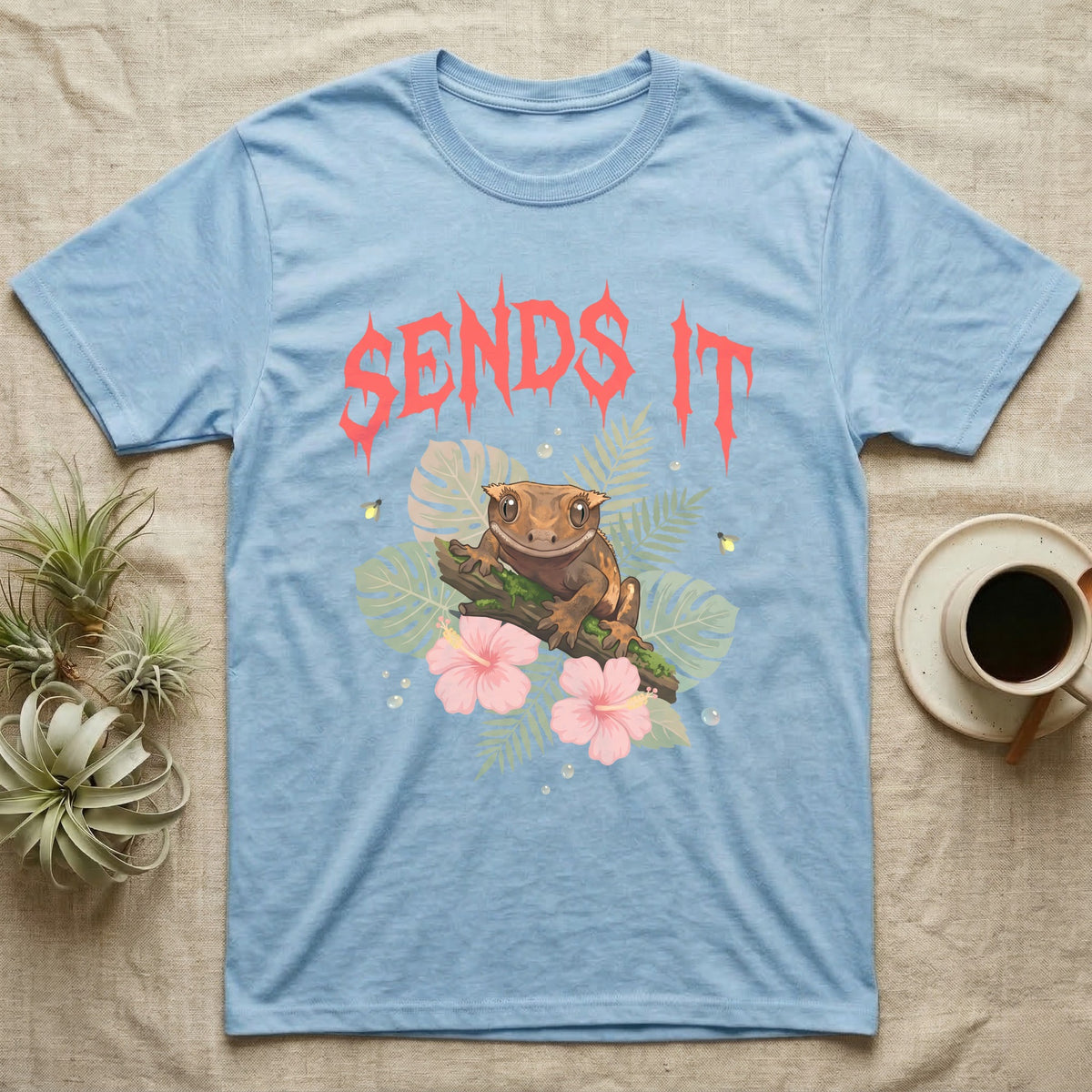 Crested Gecko Feral T-Shirt in Light Blue — Crested gecko on a mossy branch in a pastel jungle scene under a spiky coral metal wordmark reading SENDS IT.