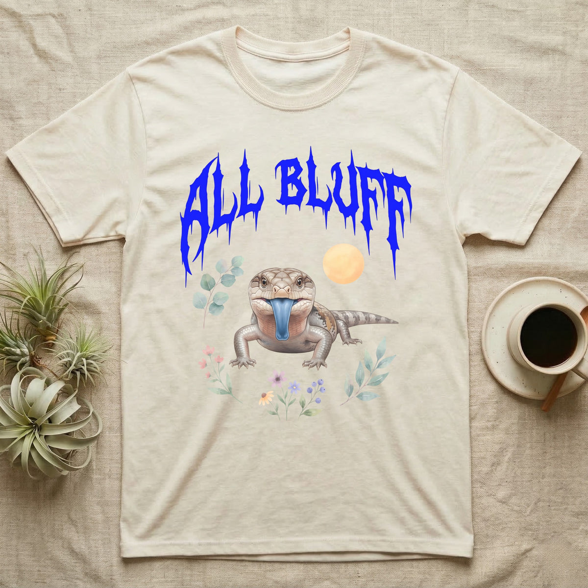 Northern Blue-Tongue Skink Feral T-Shirt in Natural — Blue-tongue skink with its blue tongue out under a jagged cobalt-blue metal "ALL BLUFF" wordmark and pastel leaves.