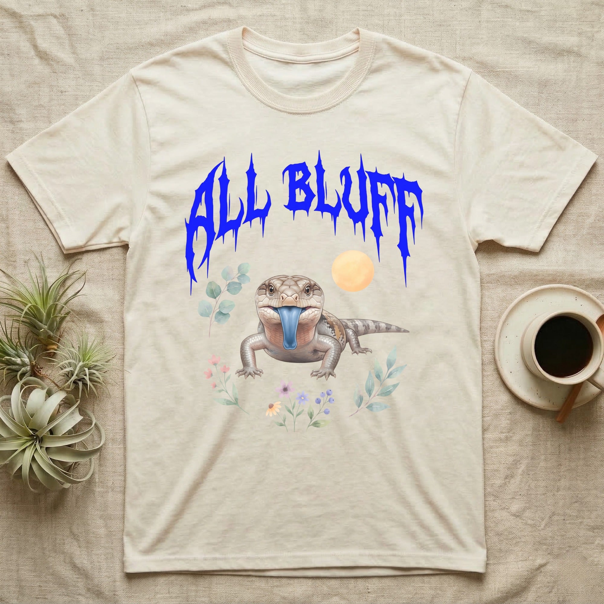 Northern Blue-Tongue Skink Feral T-Shirt in Natural — Blue-tongue skink with its blue tongue out under a jagged cobalt-blue metal "ALL BLUFF" wordmark and pastel leaves.