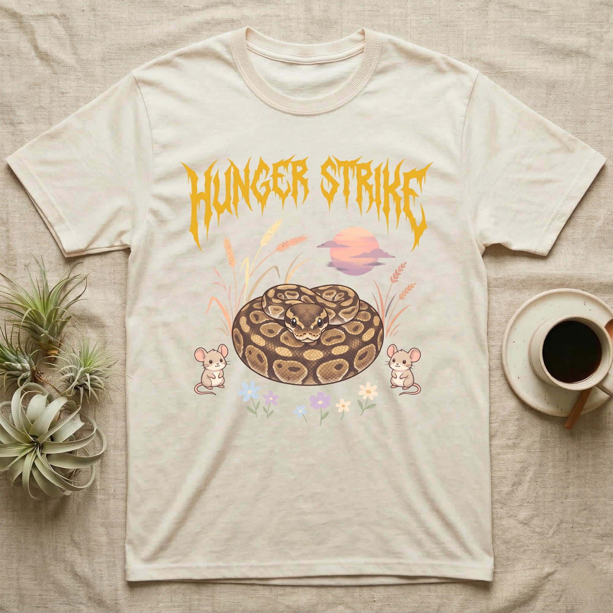 Ball Python Feral T-Shirt in Natural — Ball python coiled in a loaf in a pastel savanna scene under a spiky honey-gold metal wordmark reading HUNGER STRIKE.