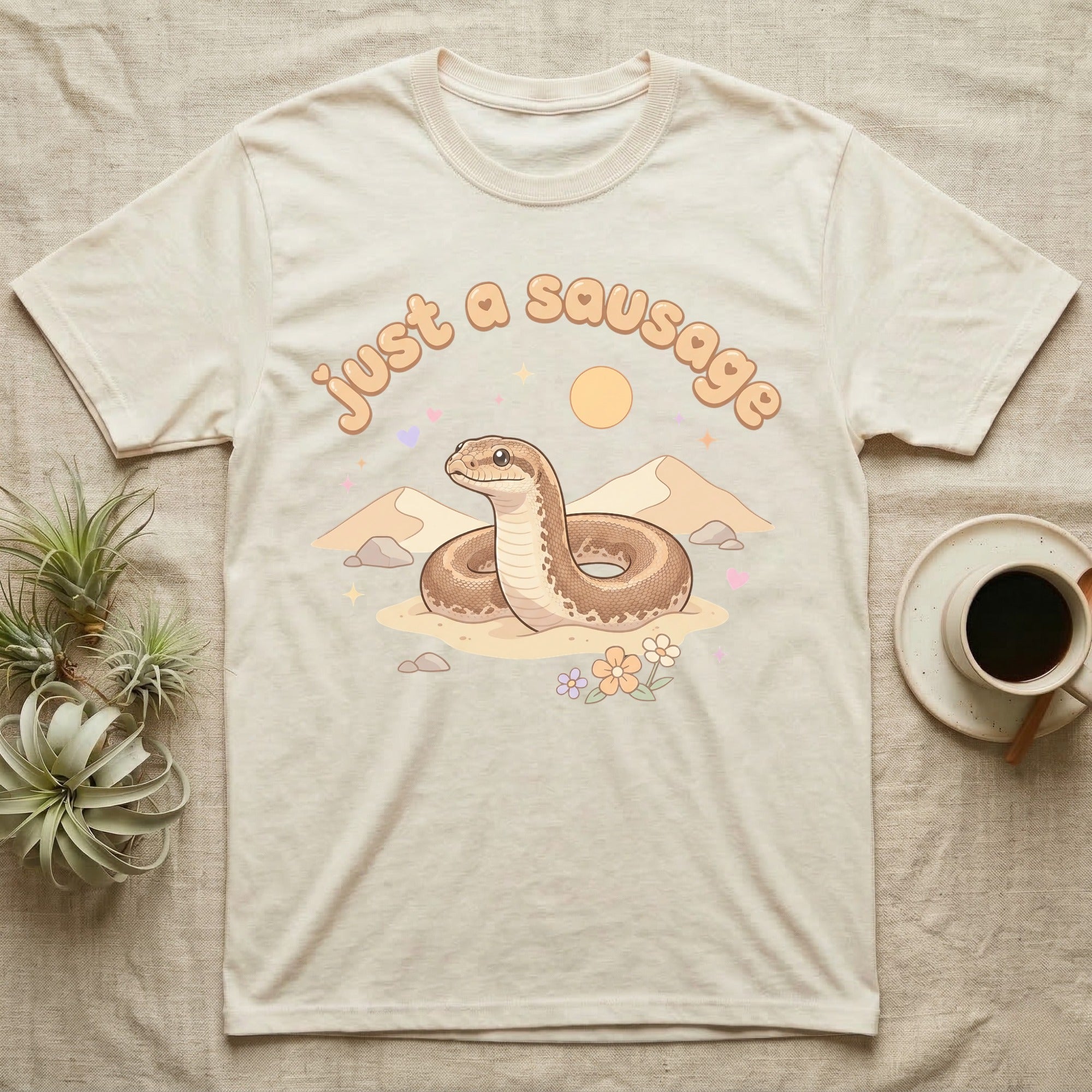 Kenyan Sand Boa Softie T-Shirt in Natural — Kenyan sand boa looping out of pastel sand dunes under a puffy "just a sausage" bubble wordmark with tiny hearts.