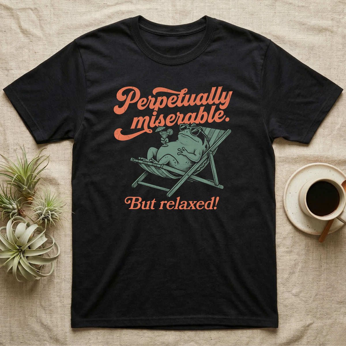 Desert Rain Frog Grump Mode T-Shirt in Black — Illustrated desert rain frog lounging in a beach chair with sunglasses and a cocktail, coral script reading Perpetually Miserable But Relaxed.
