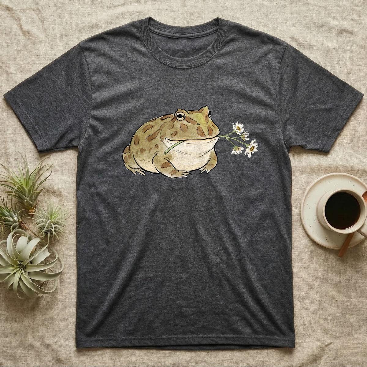 Pacman Frog Blossom T-Shirt in Dark Heather — Watercolor illustration of an olive-tan Pacman frog holding a water-lily blossom sprig, on a dark grey t-shirt.