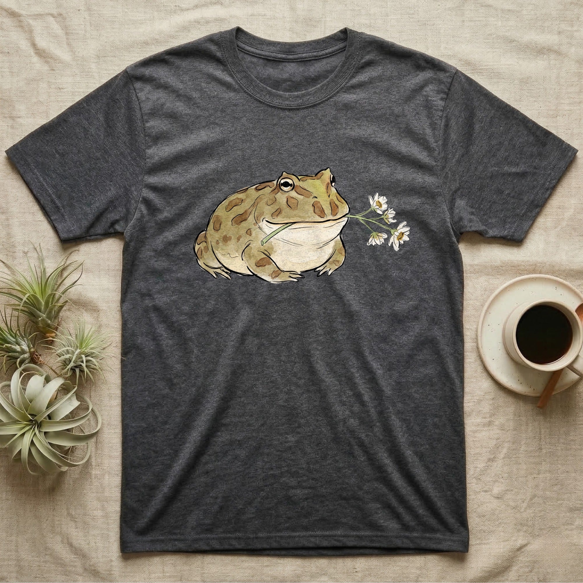 Pacman Frog Blossom T-Shirt in Dark Heather — Watercolor illustration of an olive-tan Pacman frog holding a water-lily blossom sprig, on a dark grey t-shirt.