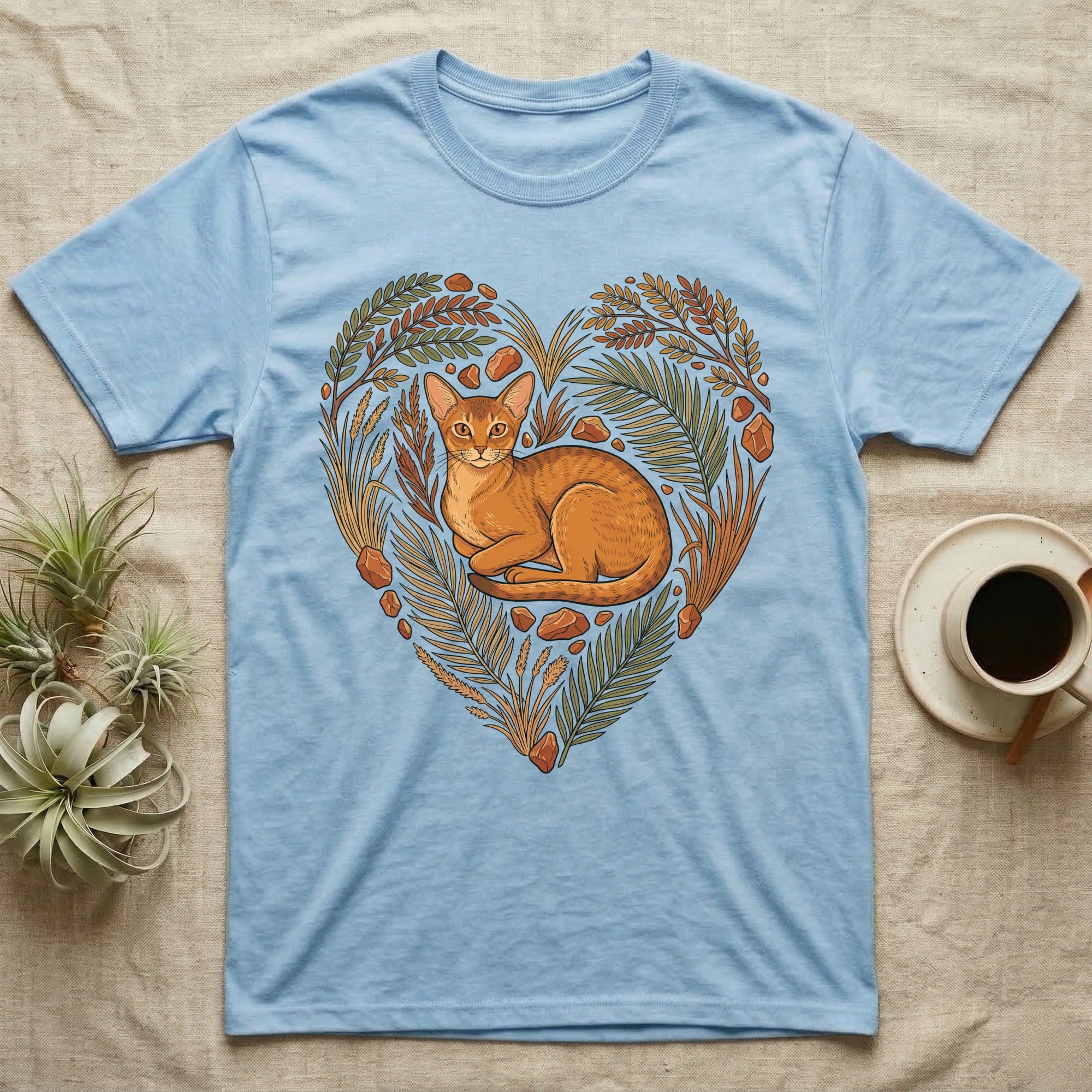 Light blue t-shirt with a heart-shaped design featuring a cat surrounded by leaves and stones.