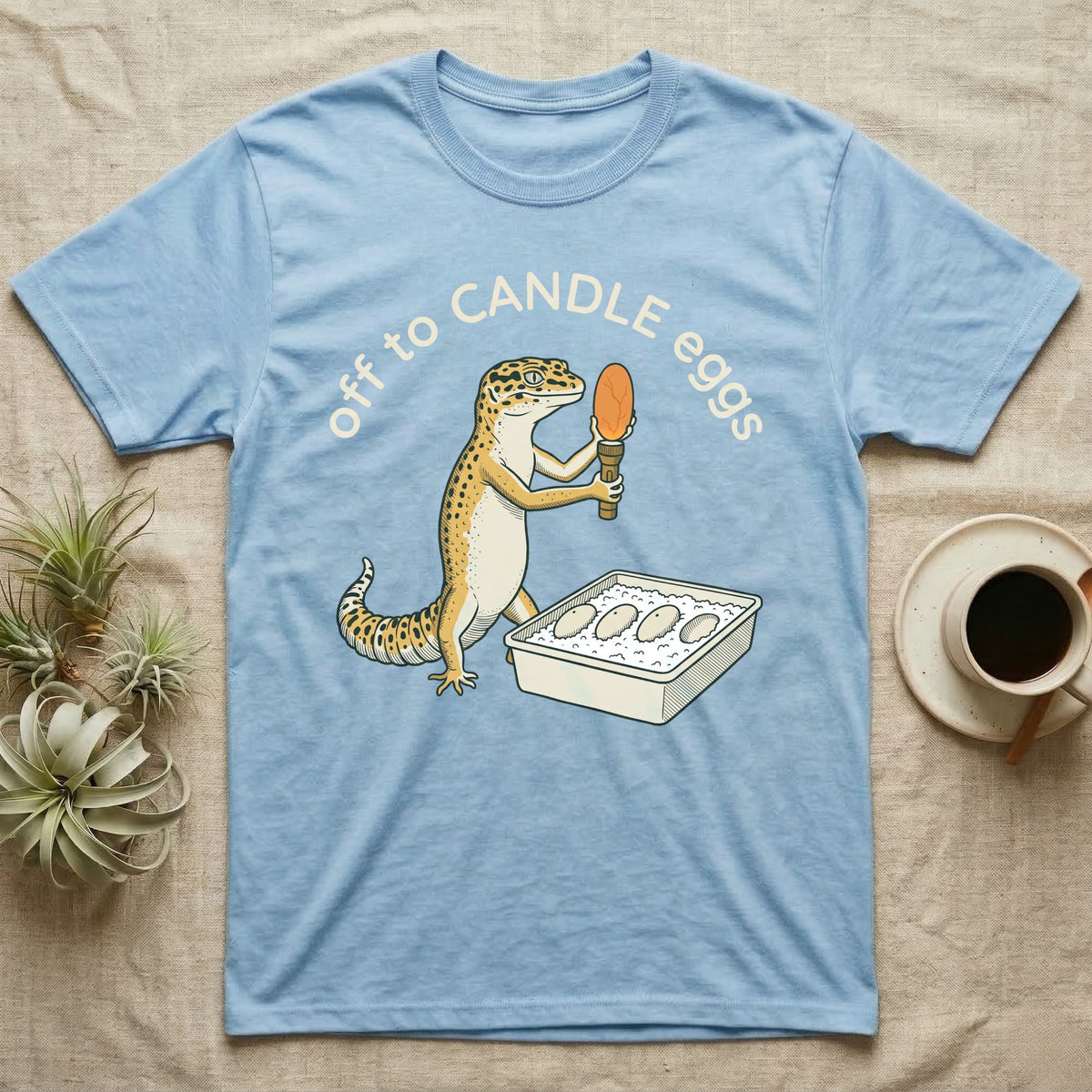 Leopard Gecko Candling T-Shirt in Light Blue — Leopard gecko holding a torch under a glowing orange egg beside a tub of eggs, under the words off to CANDLE eggs