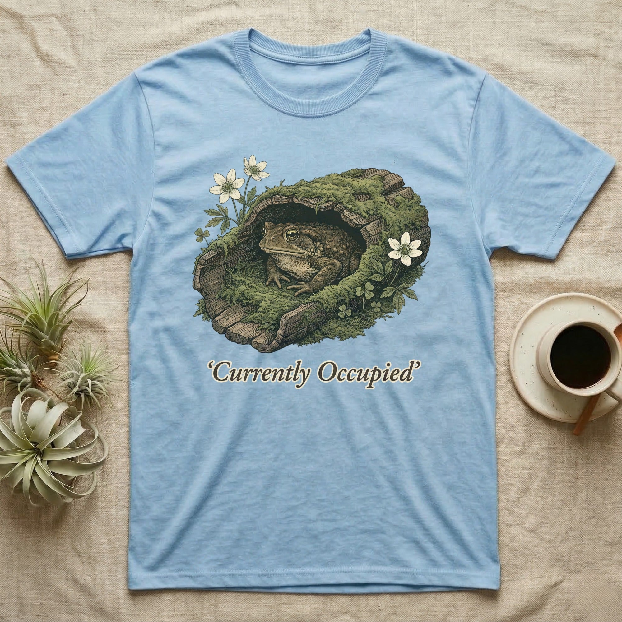 Light blue t-shirt with a toad in a mossy log with flowers, text "Currently Occupied".