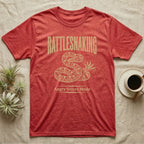 Coiled corn snake mid-tail-vibration in warm tan ink engraving beneath an arched wordmark reading RATTLESNAKING and the subtitle Angry Velcro Mode. — shown on a Heather Red tee.