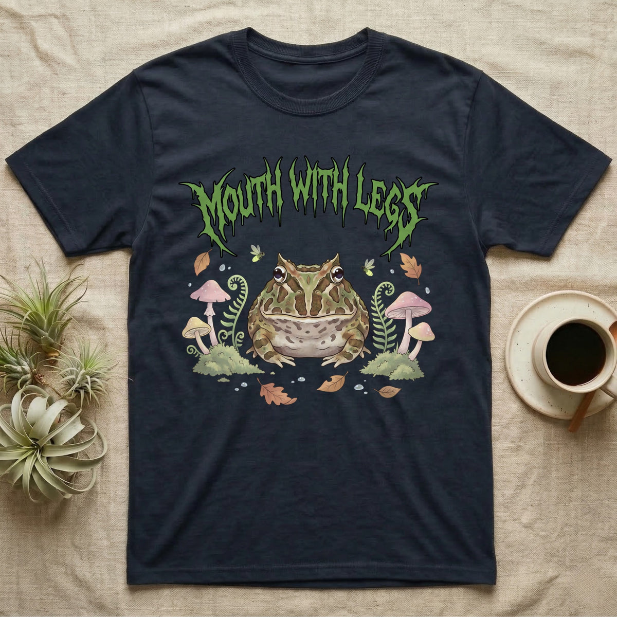 Pacman Frog Feral T-Shirt in Navy — Round Pacman frog in a pastel forest-floor scene under a spiky leaf-green metal wordmark reading MOUTH WITH LEGS.