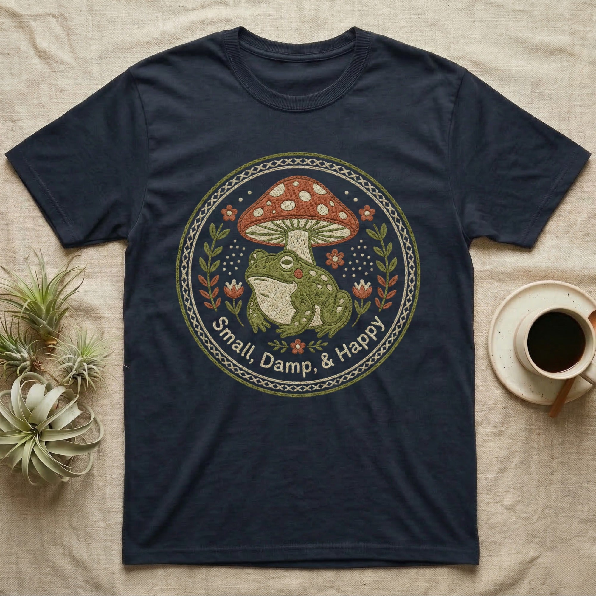 Dark t-shirt with embroidered circular design of frog, mushroom, and floral elements.