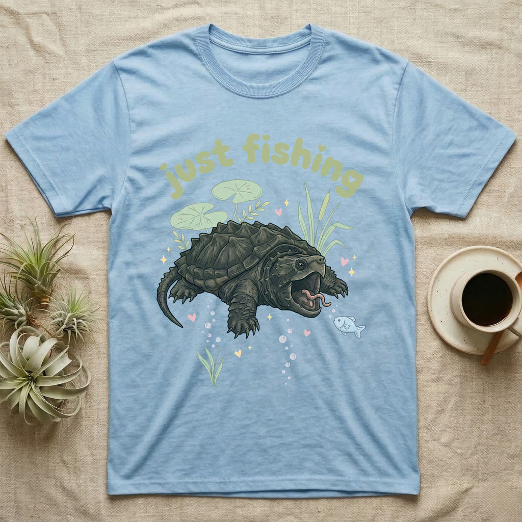 Alligator Snapping Turtle Softie T-Shirt in Light Blue — Alligator snapping turtle with open mouth and tongue lure under a puffy pastel "just fishing" bubble wordmark.