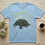 Alligator Snapping Turtle Softie T-Shirt in Light Blue — Alligator snapping turtle with open mouth and tongue lure under a puffy pastel "just fishing" bubble wordmark.