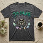 Regal Jumping Spider Feral T-Shirt in Dark Heather — Fuzzy jumping spider with huge eyes in a pastel garden under a spiky iridescent-teal metal wordmark reading TINY STALKER.