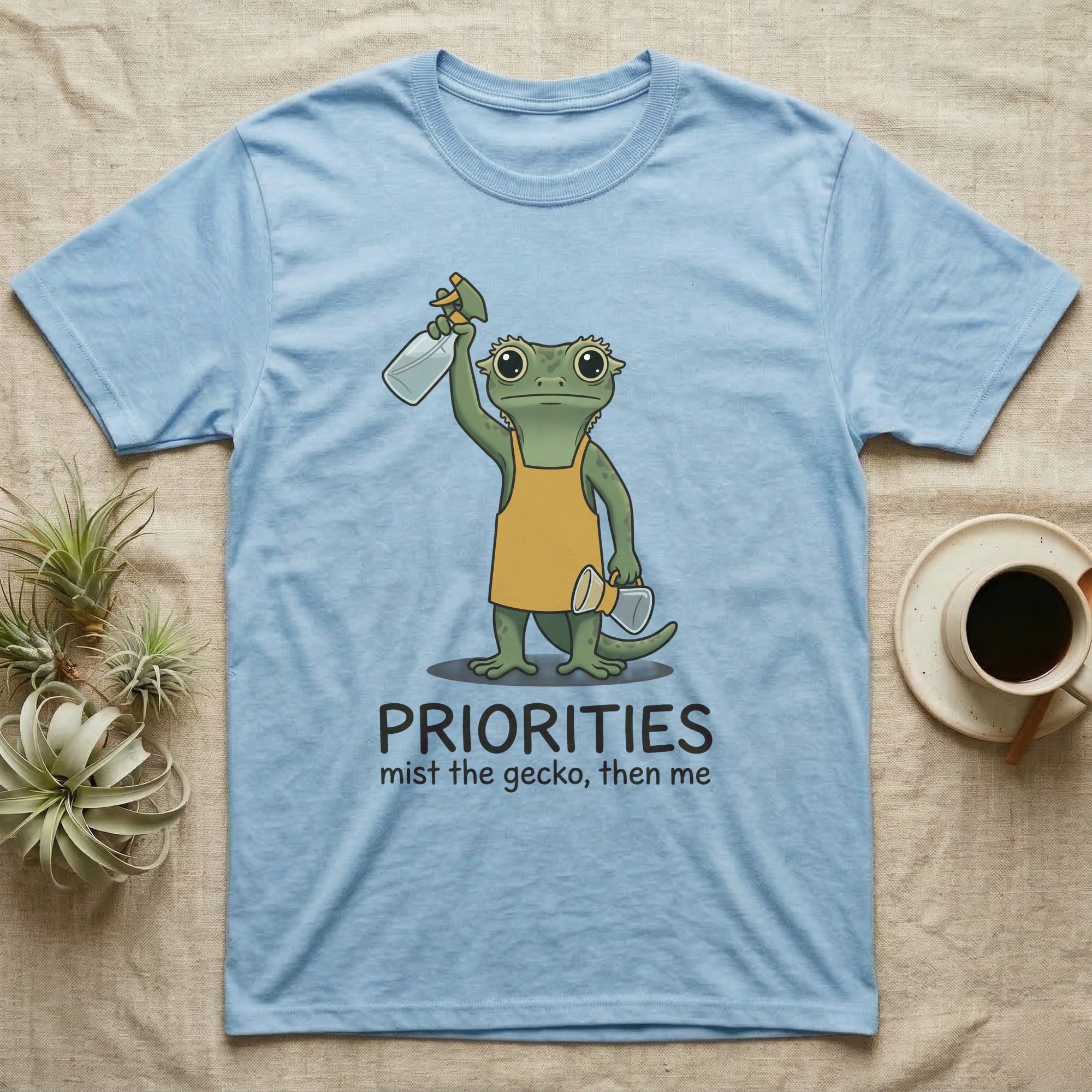 Crested Gecko Coffee T-Shirt in Light Blue — Storybook crested gecko standing with a raised spray mister and a pour-over coffee, text priorities below