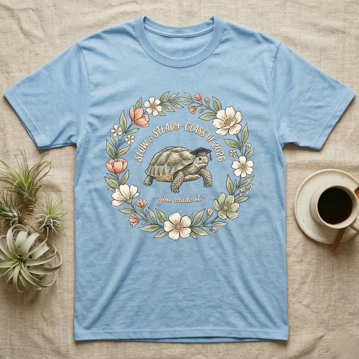 Slow And Steady Graduation T-Shirt in Light Blue — Watercolour illustration of a tortoise in a graduation cap inside a floral wreath with "Slow & Steady, Class of 2026."