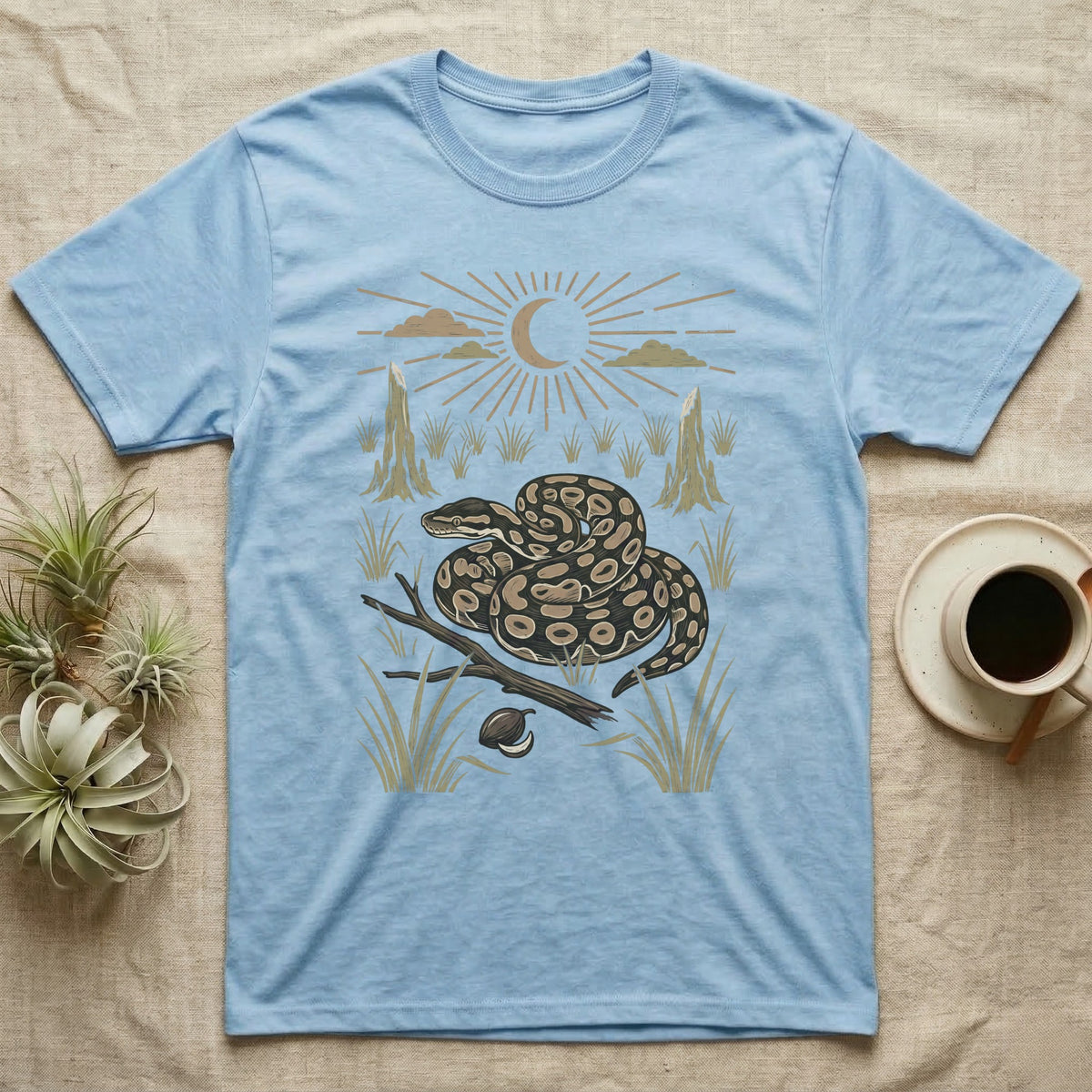 Ball Python Home Range T-Shirt in Light Blue — Woodcut-style illustration of a ball python coiled on dry savanna ground with grass tufts and termite mounds behind, no on-shirt text.