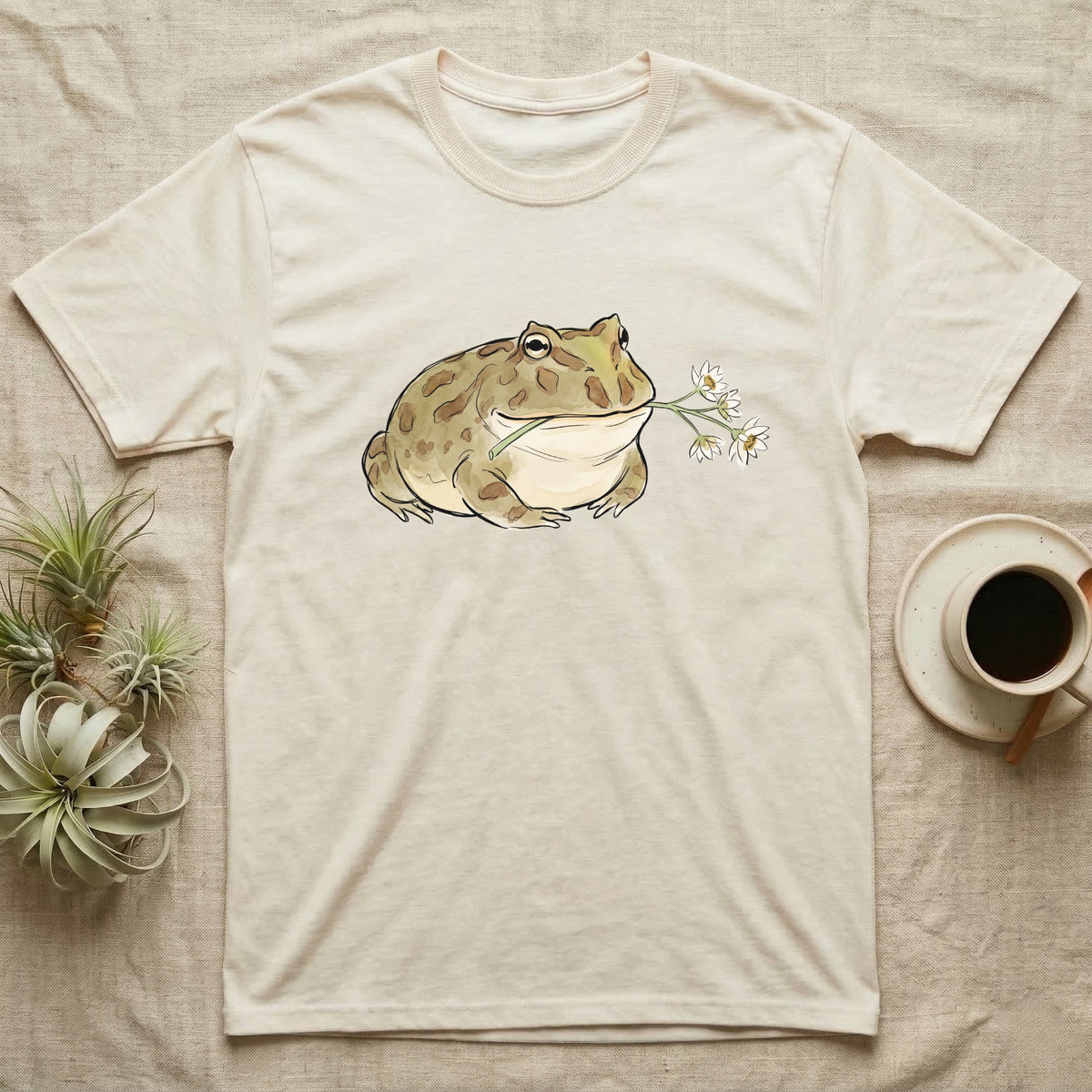 Pacman Frog Blossom T-Shirt in Natural — Watercolor illustration of an olive-tan Pacman frog holding a water-lily blossom sprig, on a dark grey t-shirt.