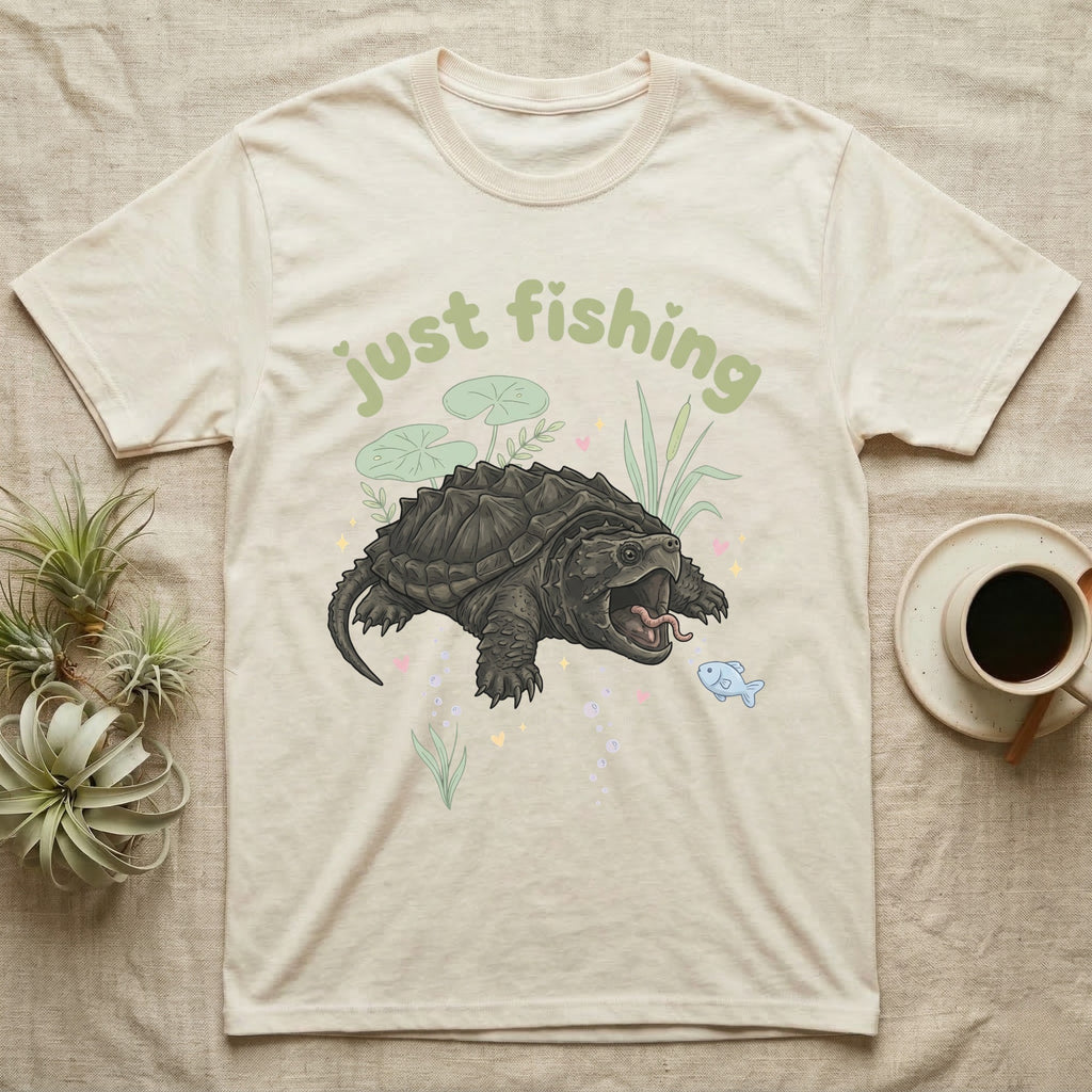 Alligator Snapping Turtle Softie T-Shirt in Natural — Alligator snapping turtle with open mouth and tongue lure under a puffy pastel "just fishing" bubble wordmark.
