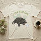 Alligator Snapping Turtle Softie T-Shirt in Natural — Alligator snapping turtle with open mouth and tongue lure under a puffy pastel "just fishing" bubble wordmark.