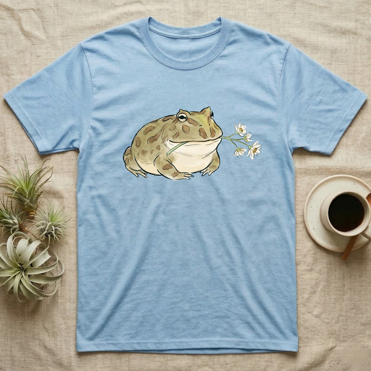 Pacman Frog Blossom T-Shirt in Light Blue — Watercolor illustration of an olive-tan Pacman frog holding a water-lily blossom sprig, on a dark grey t-shirt.