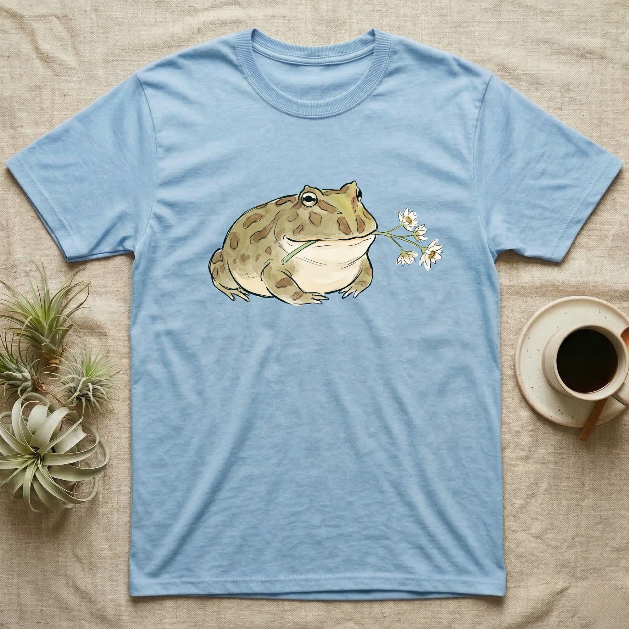 Pacman Frog Blossom T-Shirt in Light Blue — Watercolor illustration of an olive-tan Pacman frog holding a water-lily blossom sprig, on a dark grey t-shirt.