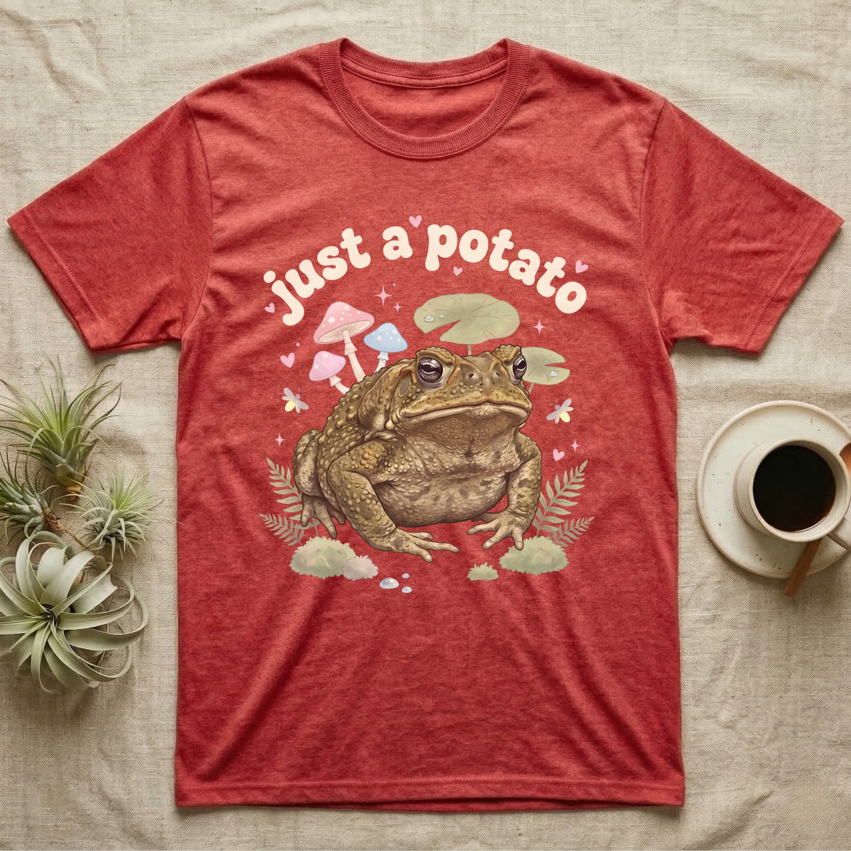 Cane Toad Baby T-Shirt in Heather Red — Warty cane toad among pastel toadstools and lily pads under puffy cream bubble text "just a potato".
