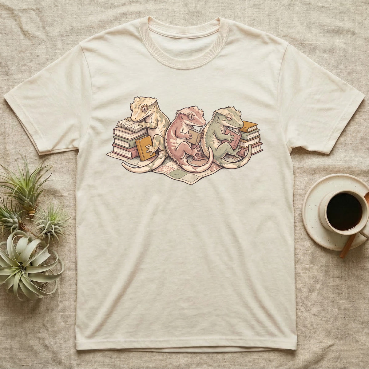 Gecko Book Club T-Shirt in Natural — Warm storybook illustration of three crested geckos in a cozy reading row, each beside a tiny stack of books.