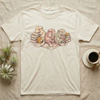 Gecko Book Club T-Shirt in Natural — Warm storybook illustration of three crested geckos in a cozy reading row, each beside a tiny stack of books.