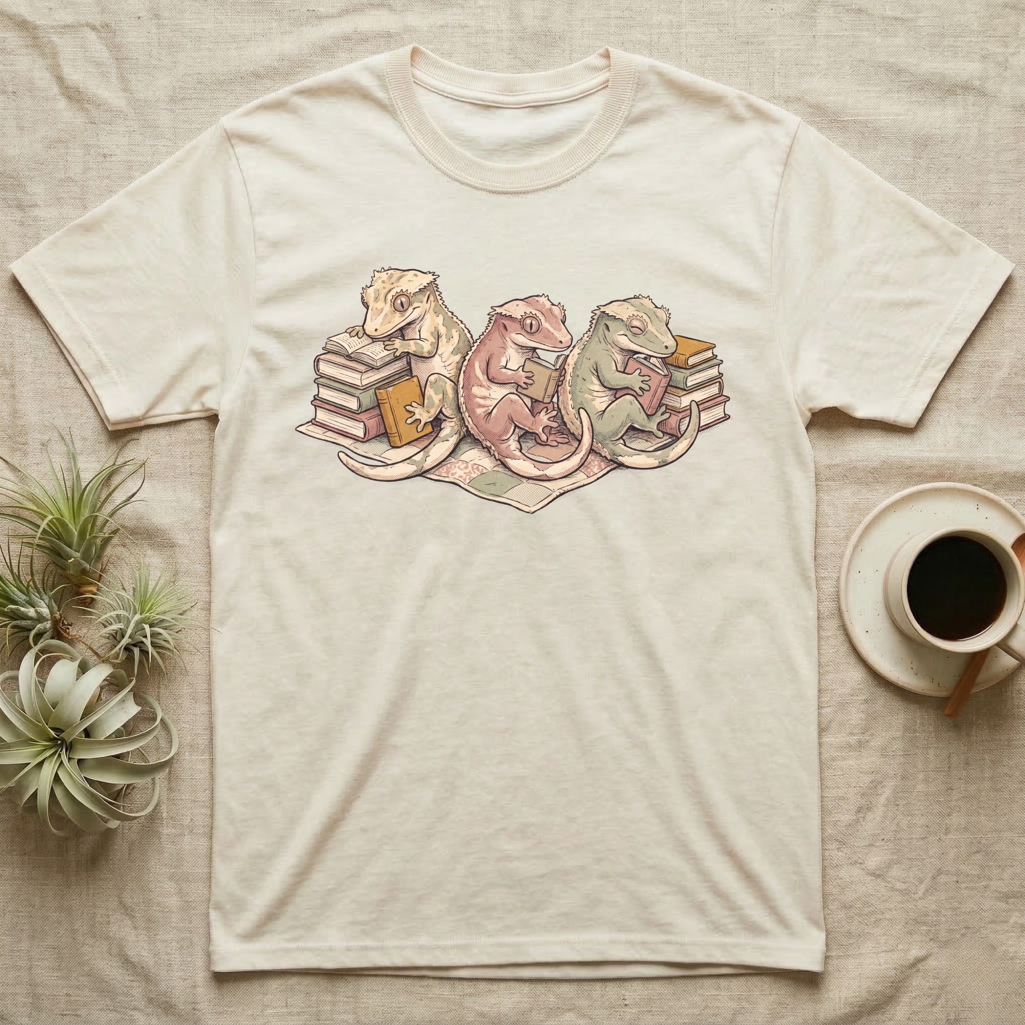 Gecko Book Club T-Shirt in Natural — Warm storybook illustration of three crested geckos in a cozy reading row, each beside a tiny stack of books.