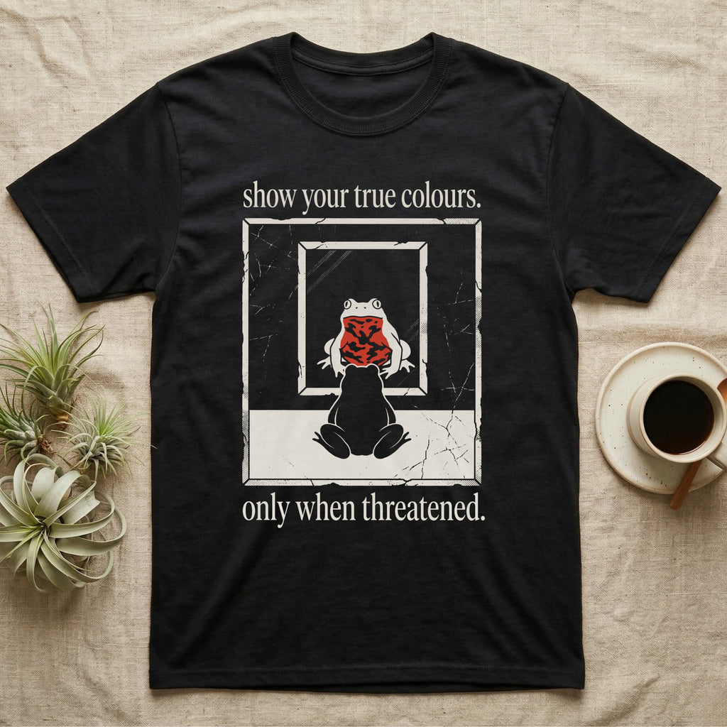 Fire-Bellied Toad Mirror T-Shirt in Black — Worn photocopy-style poster of a fire-bellied toad seen from behind looking into a mirror, its reflection showing a bright red marbled belly, with the words show your true colours, only when threatened.