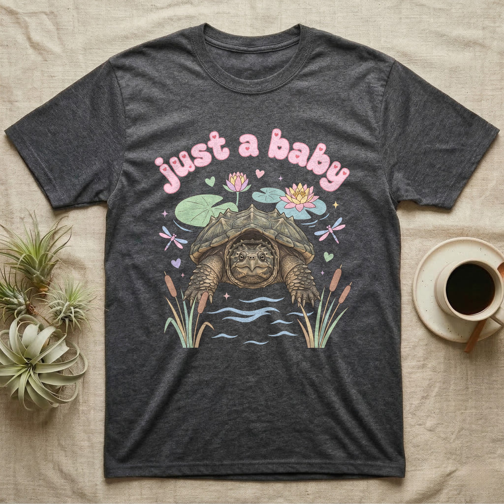 Common Snapping Turtle Baby T-Shirt in Dark Heather — Snapping turtle with hooked beak and spiky shell among pastel lily pads under puffy pink bubble text "just a baby".