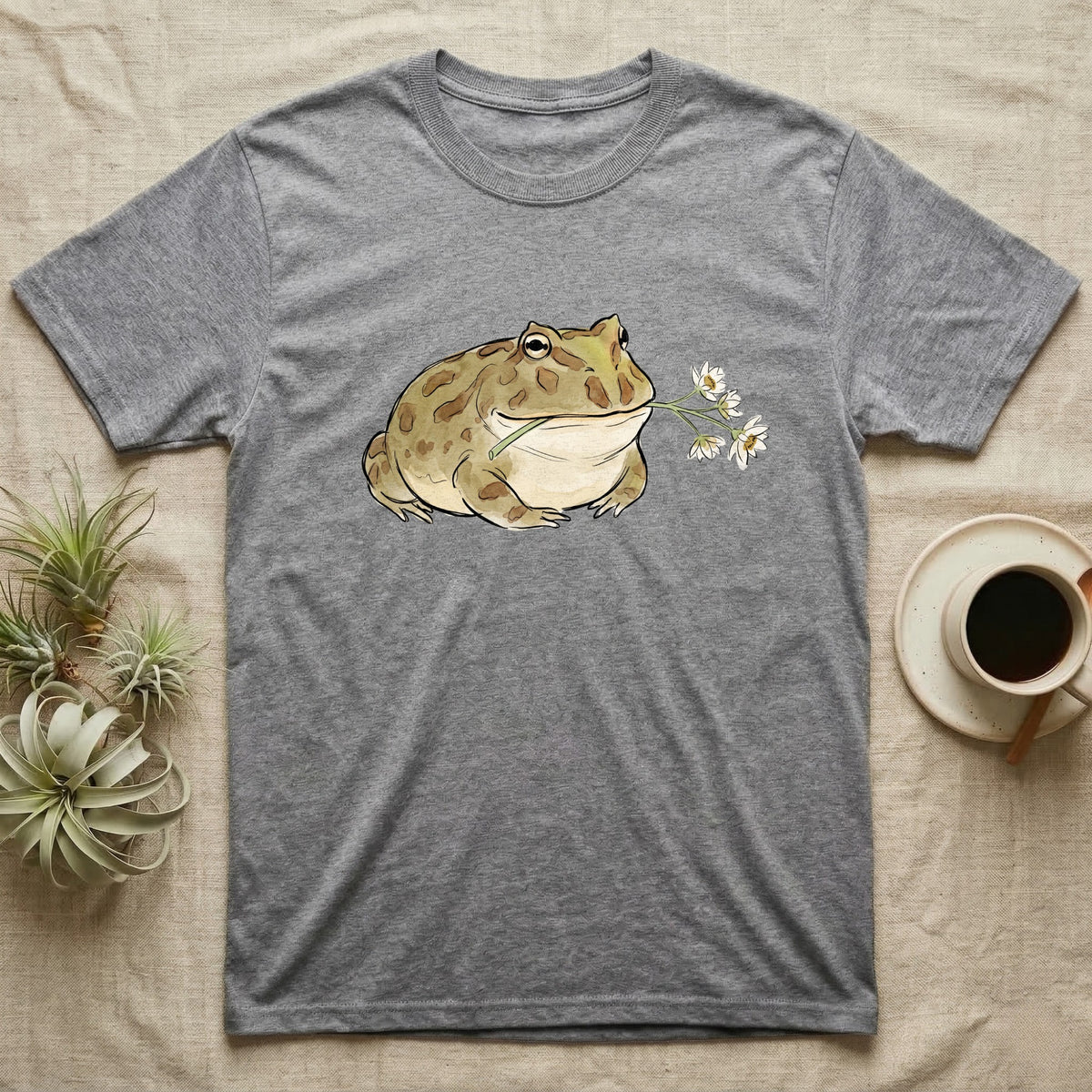 Pacman Frog Blossom T-Shirt in Sport Grey — Watercolor illustration of an olive-tan Pacman frog holding a water-lily blossom sprig, on a dark grey t-shirt.