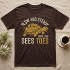 Hermann's Tortoise Toes T-Shirt in Dark Chocolate — Golden ochre Hermann's tortoise charging to the right under arched cream words Slow And Steady, with Until She Sees Toes below