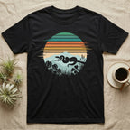 Ball Python Retro Sunset T-Shirt in Black — Ball python silhouette draped over a branch inside a retro striped sunset disc with monstera leaves on a tee