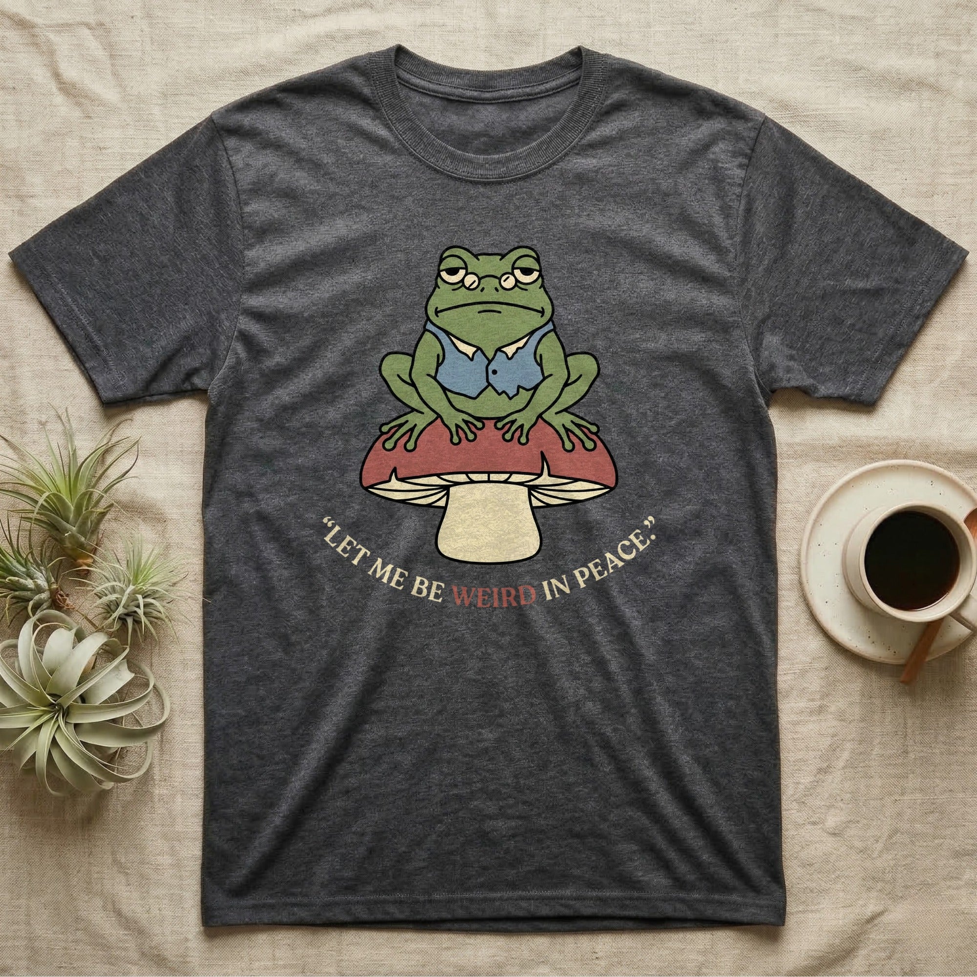 Dark grey t-shirt with a frog on a mushroom and the text "Let me be weird in peace."